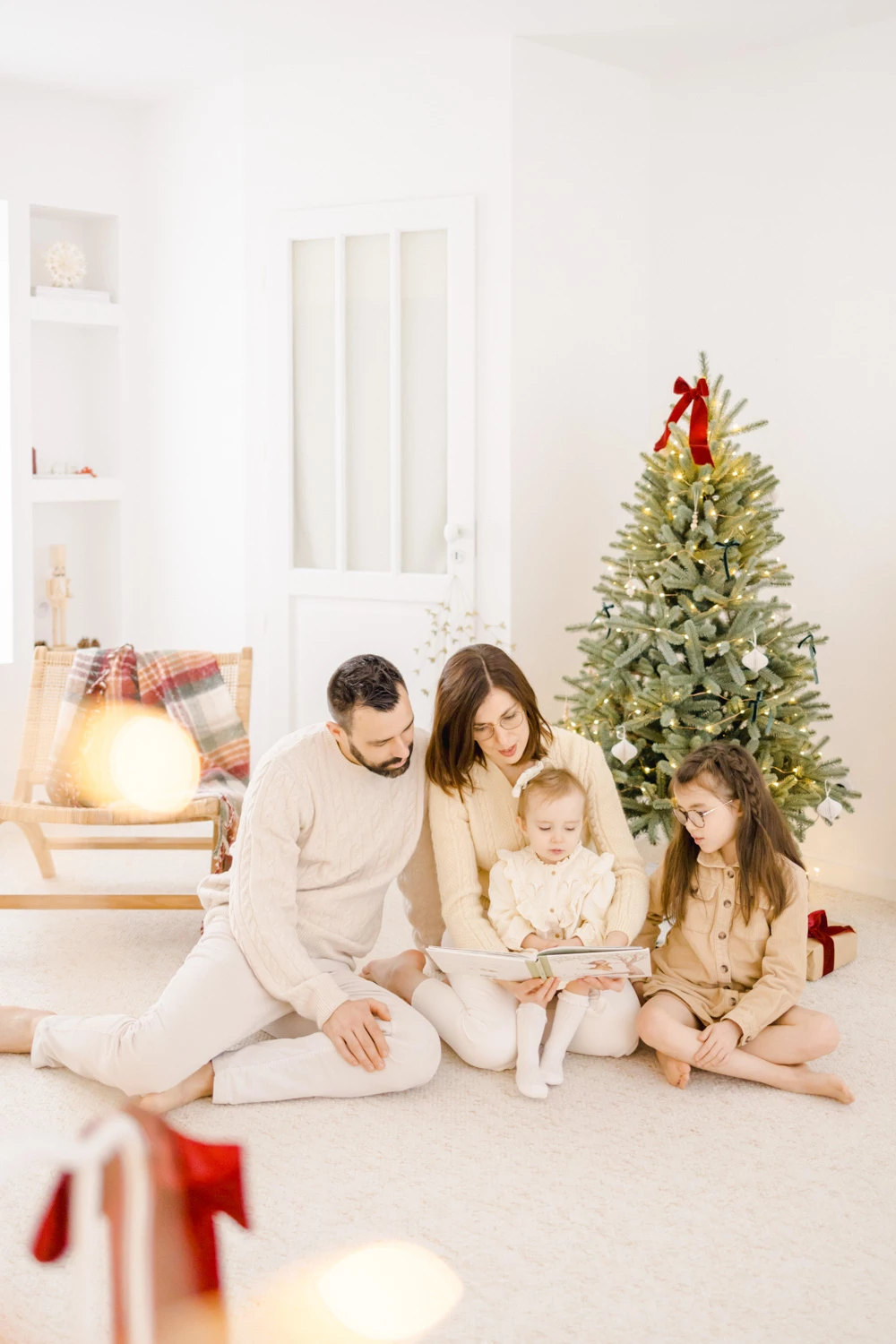 Séance photo famille Noël : parents et deux enfants en tenues beiges assis près du sapin, ambiance chaleureuse et intime en lumière naturelle douce