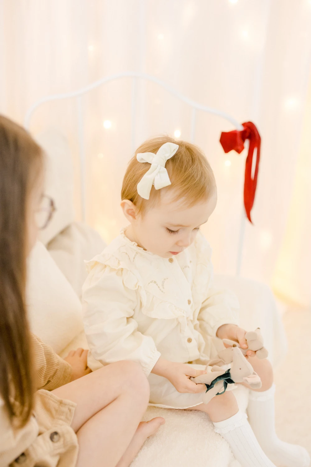 Séance photo famille nouveau-né : petit enfant en tenue blanche avec noeud beige regardant un jouet, dans une ambiance lumineuse et chaleureuse