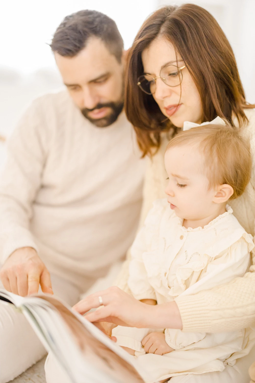 Séance famille intemporelle : parents et enfant en tenues beige clair partageant un moment de complicité autour d'un livre, lumière douce et naturelle, ambiance chaleureuse et intimiste