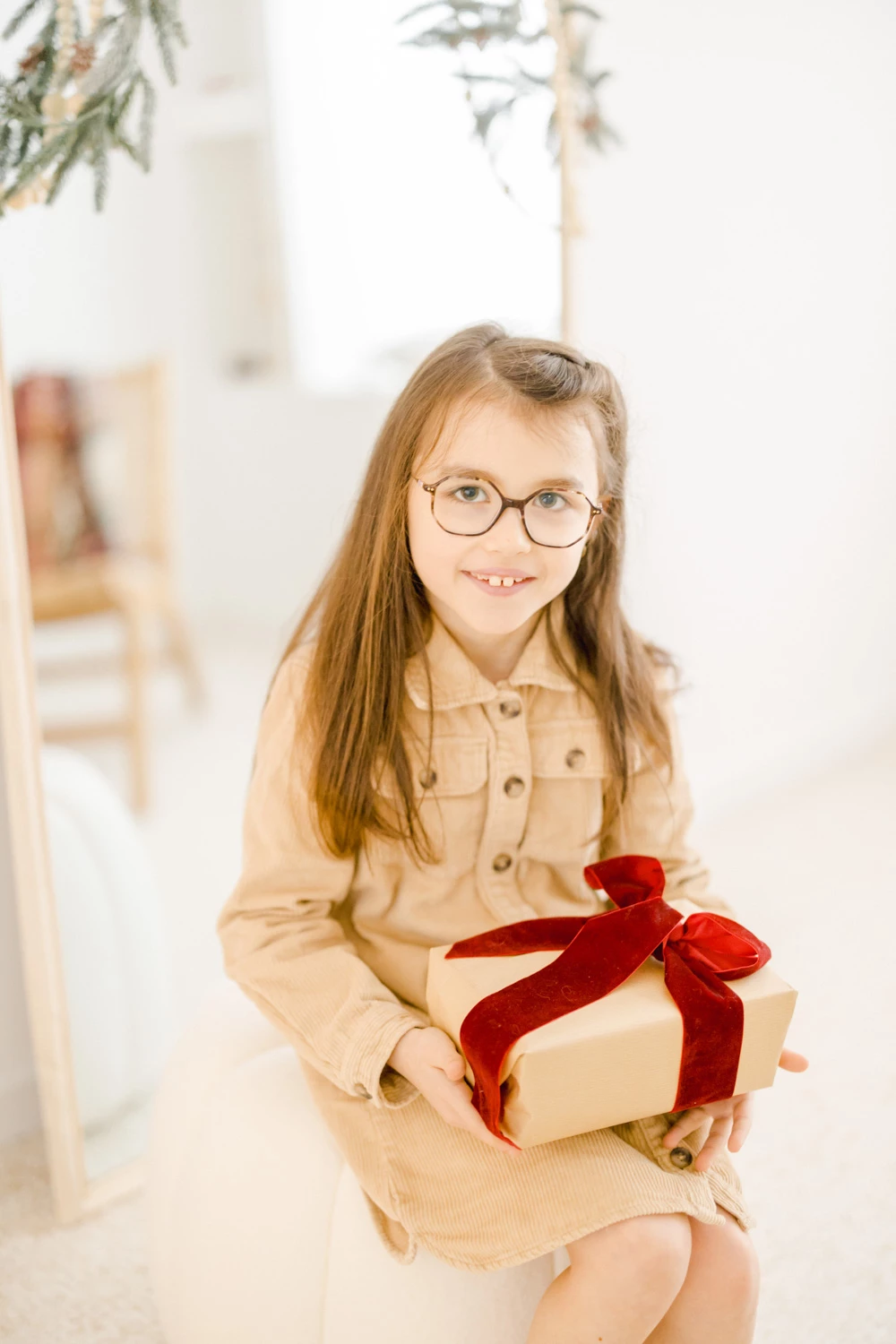 Portrait d'une jeune fille souriante portant des lunettes et une chemise beige clair, tenant un cadeau enrubanné de rouge dans un intérieur lumineux et épuré