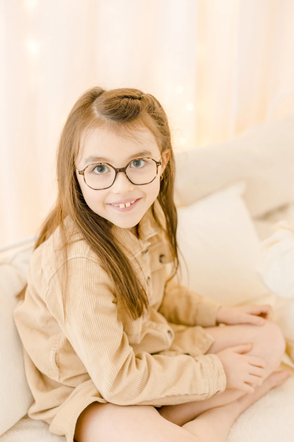 Portrait enfant fille aux cheveux roux portant des lunettes, souriante en chemise beige clair, assis sur fond blanc lumineux en studio