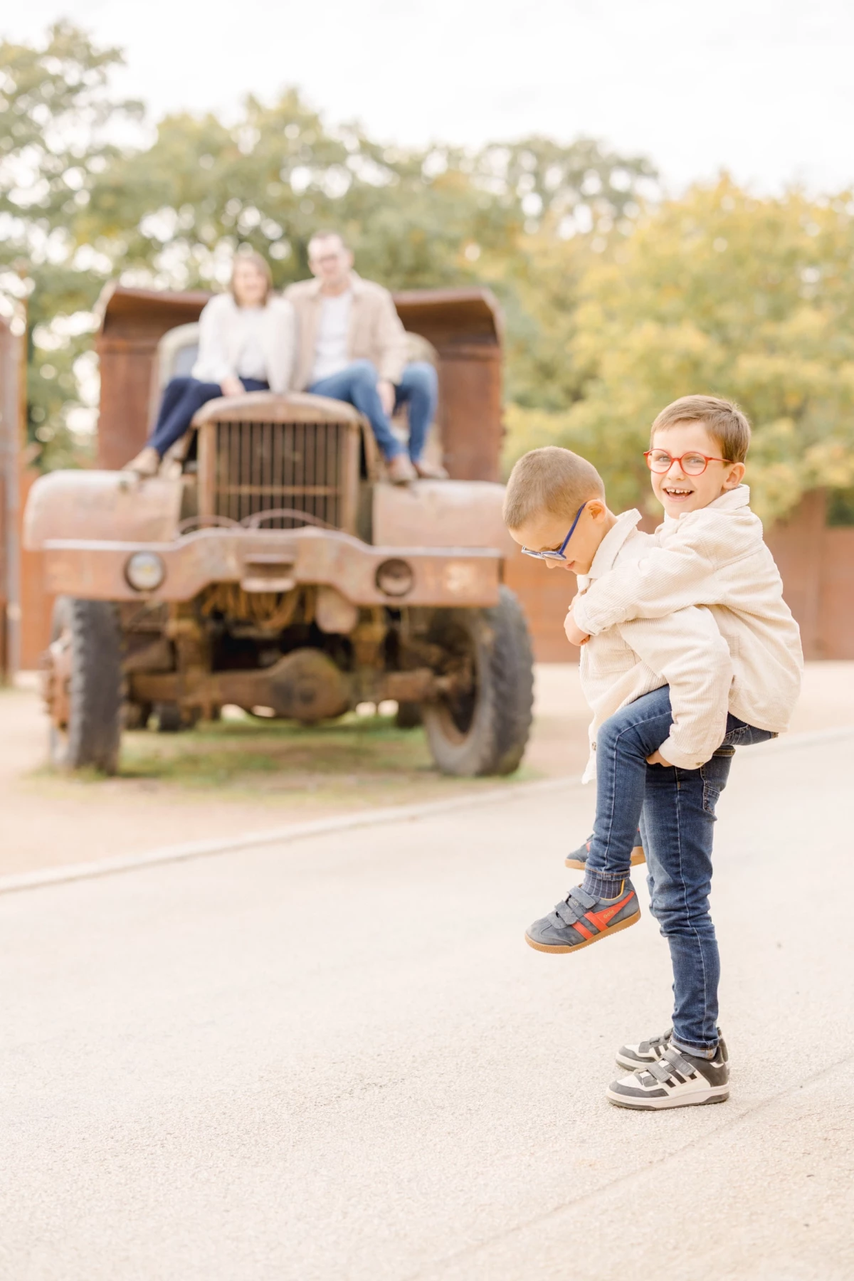 Deux enfants complices enlacés devant un tracteur vintage, famille en arrière-plan dans la campagne