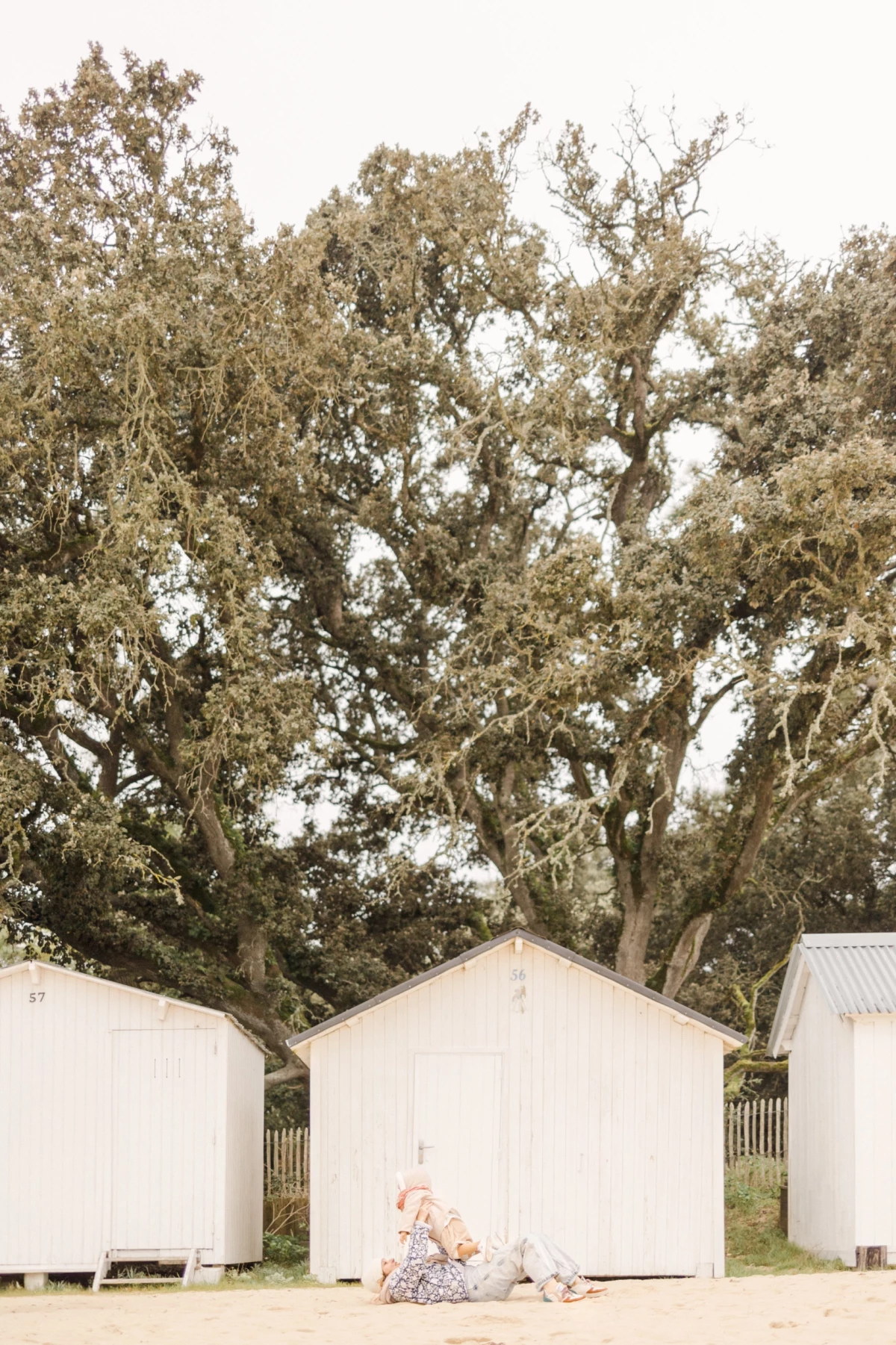 Enfant assis devant une cabane de plage blanche sous de grands arbres, ambiance estivale douce