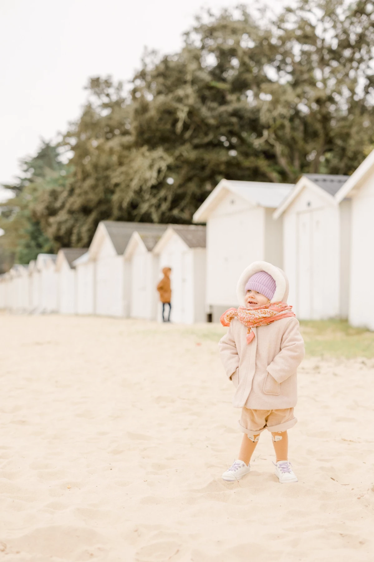 Petite fille souriante en manteau beige et écharpe orange sur une plage de sable fin