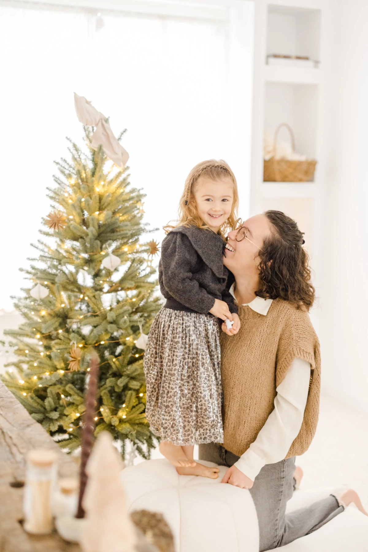 Mère et fille complices devant un sapin de Noël illuminé dans un intérieur blanc et chaleureux