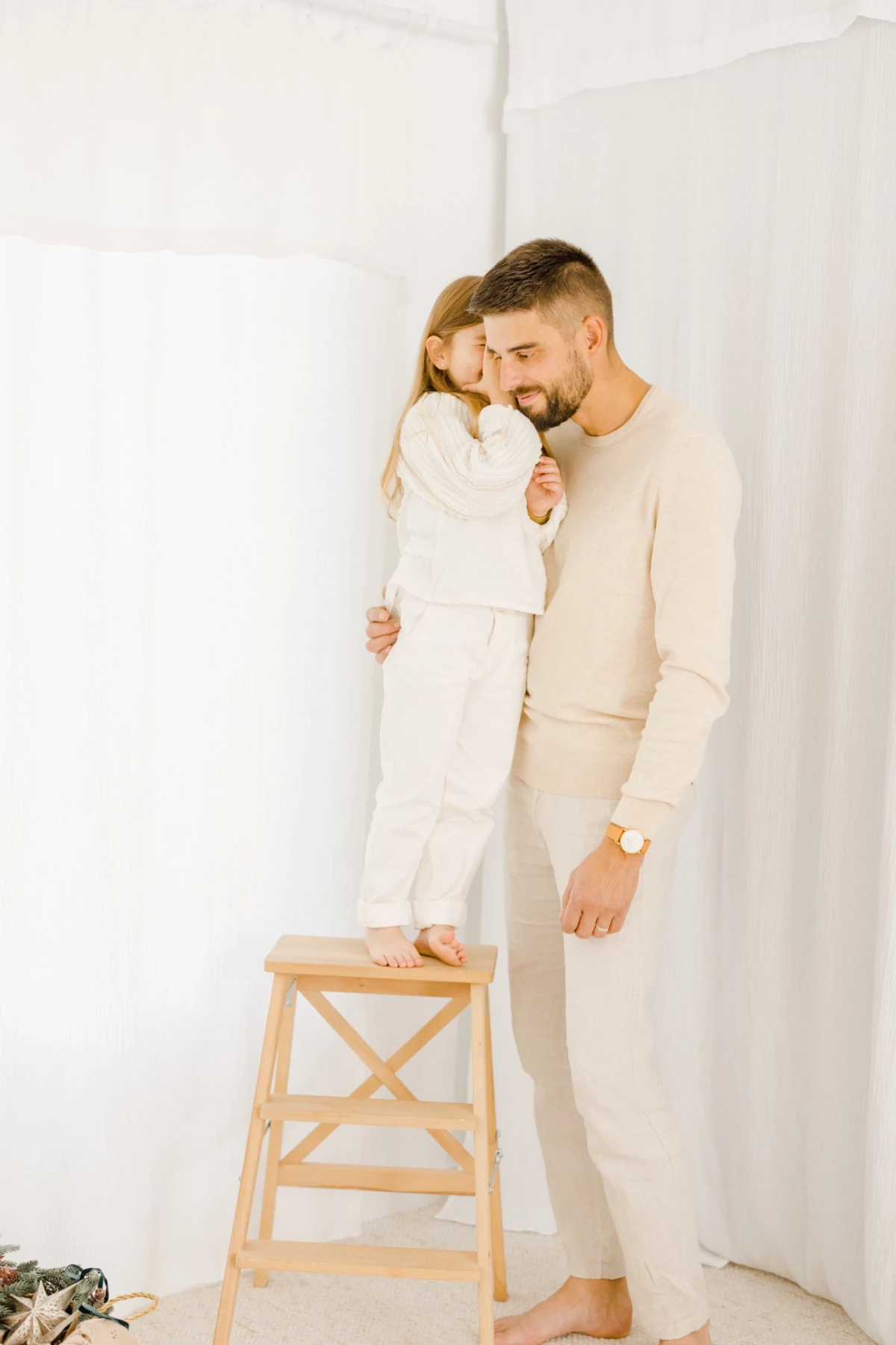 Père et fille complices en tenue beige, la fillette debout sur un tabouret enlace tendrement son papa