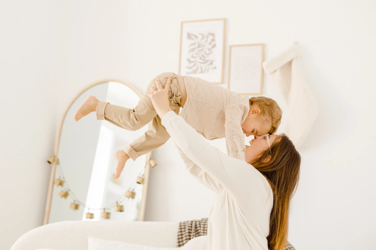 Maman soulevant son bébé dans les bras dans une chambre lumineuse aux tons blancs et beiges