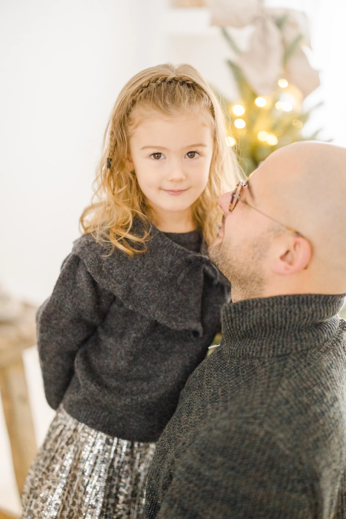 Père tenant sa petite fille en pull gris devant un sapin de Noël illuminé