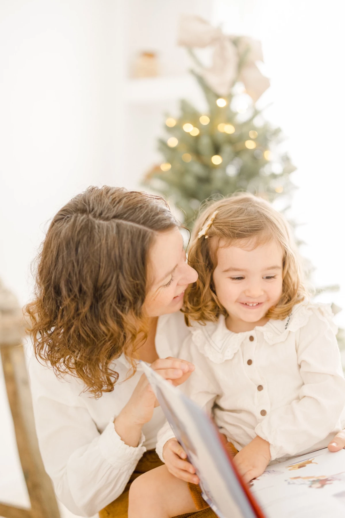 Mère et sa fille lisant un livre ensemble devant le sapin de Noël illuminé dans un intérieur blanc