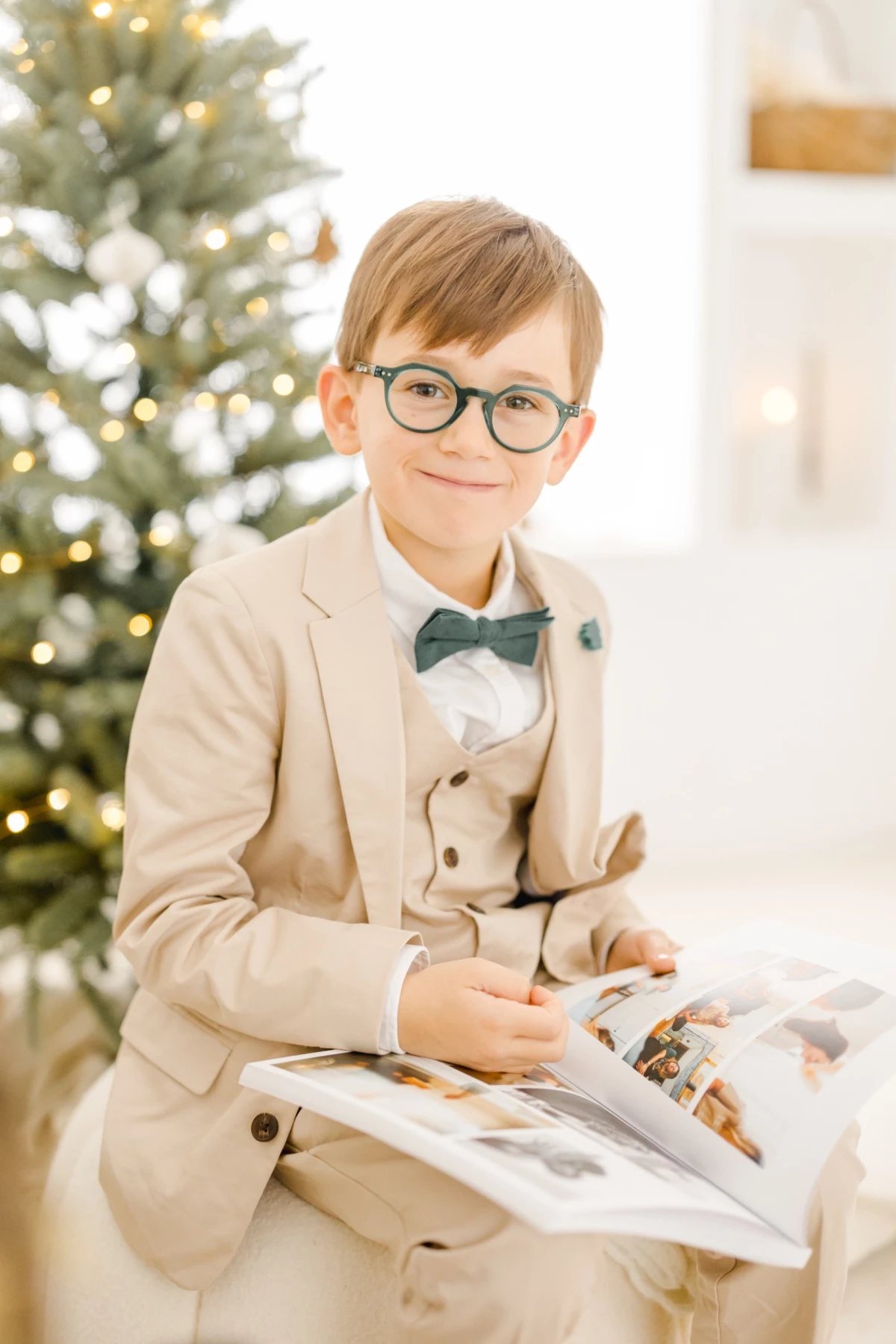 Enfant élégant en costume beige et nœud papillon, lisant près d'un sapin illuminé de Noël