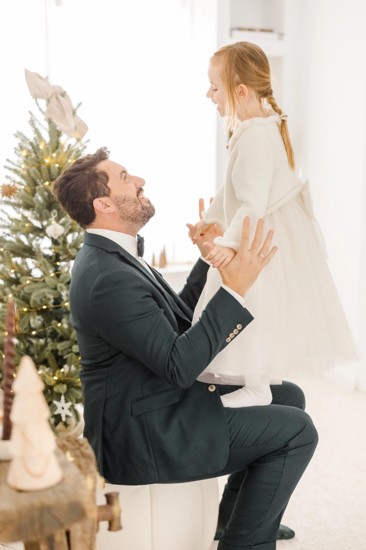 Père en costume élégant soulevant sa fille en robe blanche devant un sapin de Noël décoré