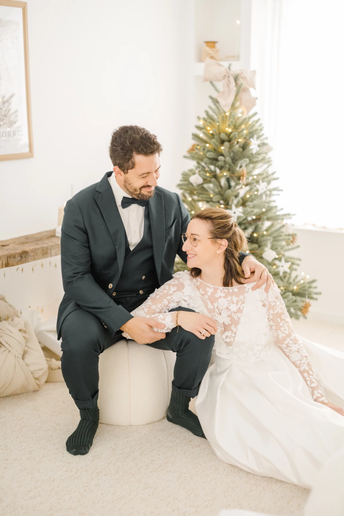 Couple de mariés assis côte à côte devant un sapin de Noël, échangeant un regard tendre et complice