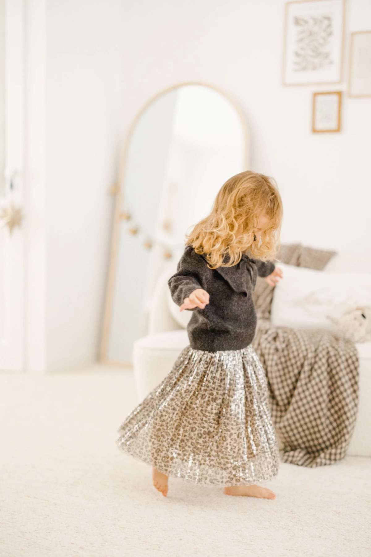 Petite fille blonde dansant pieds nus en jupe à paillettes dorées et pull gris dans une chambre lumineuse