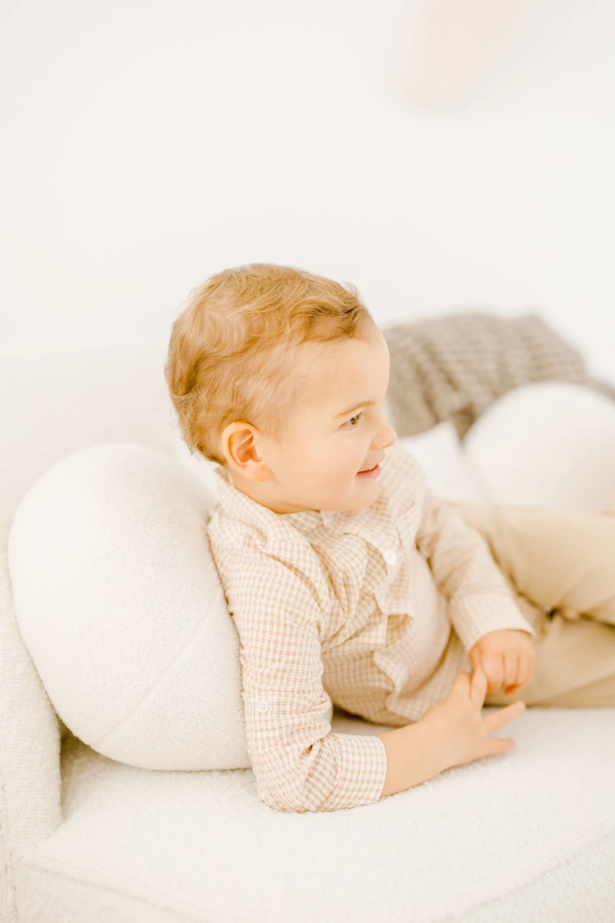 Bébé blond en tenue beige assise contre un coussin blanc en studio, regard tourné vers la lumière