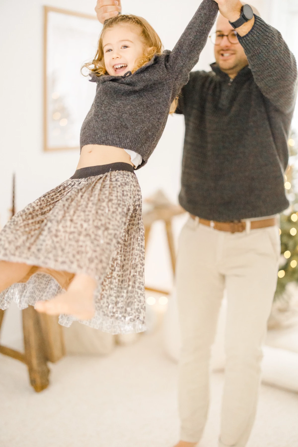 Père soulevant sa fille joyeuse en pull gris et jupe léopard dans un intérieur lumineux décoré pour Noël