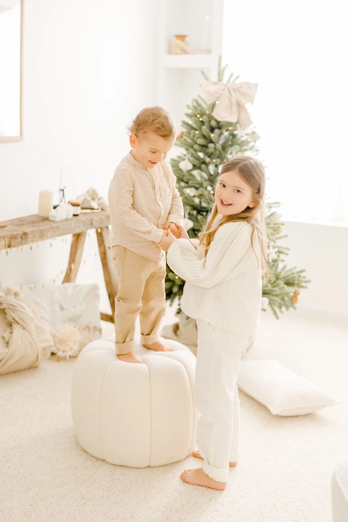 Deux enfants complices jouant près d'un sapin de Noël dans un décor blanc lumineux et chaleureux