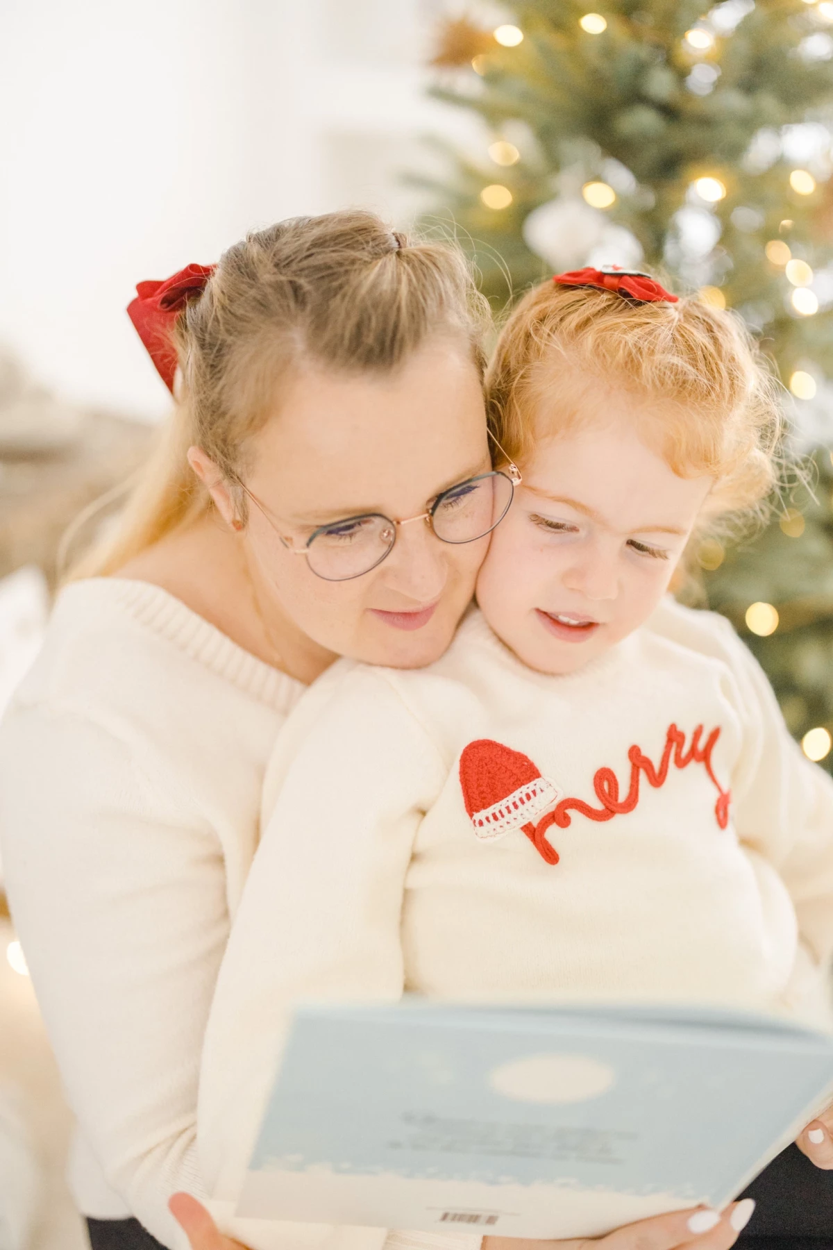 Deux fillettes blondes lisent un livre ensemble devant un sapin de Noël illuminé