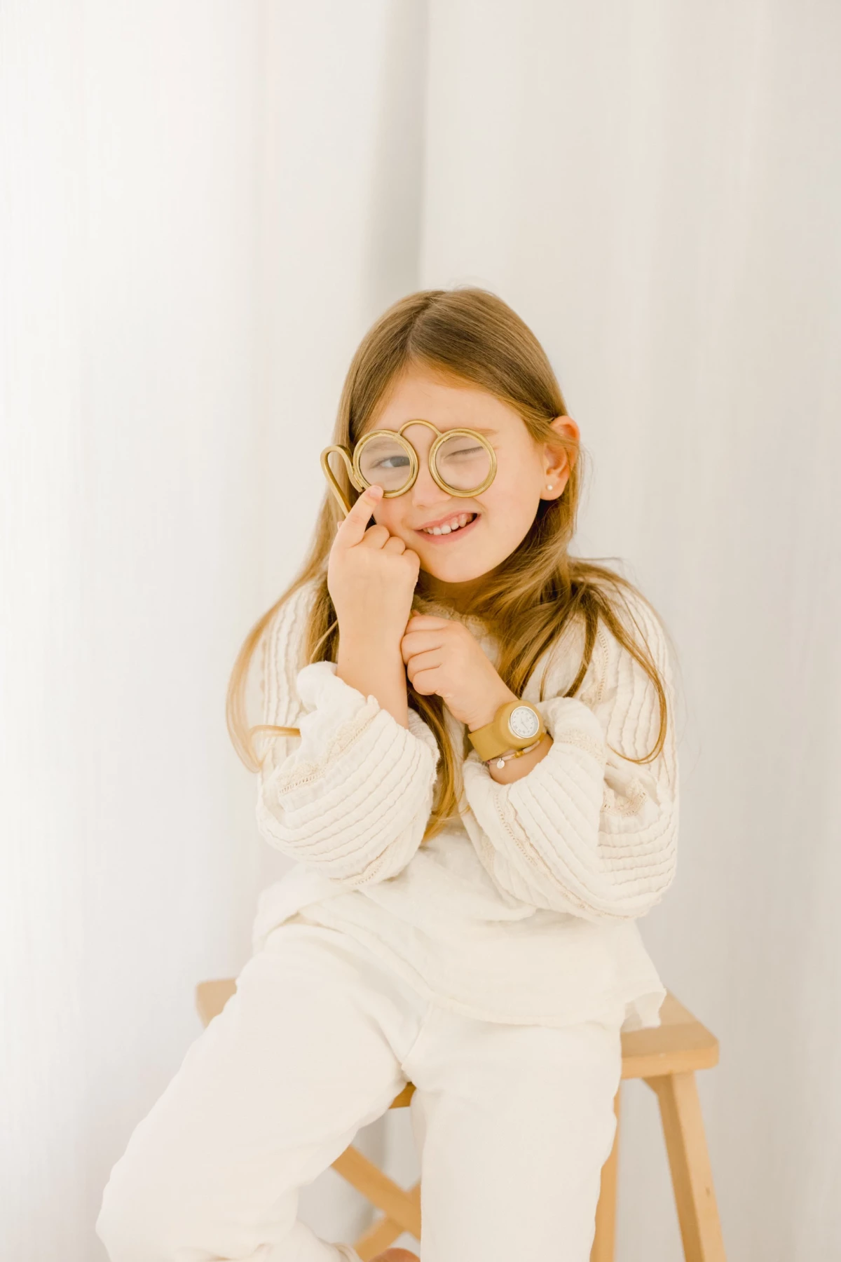 Petite fille souriante en pull blanc tricot assise sur chaise en bois, fond studio clair