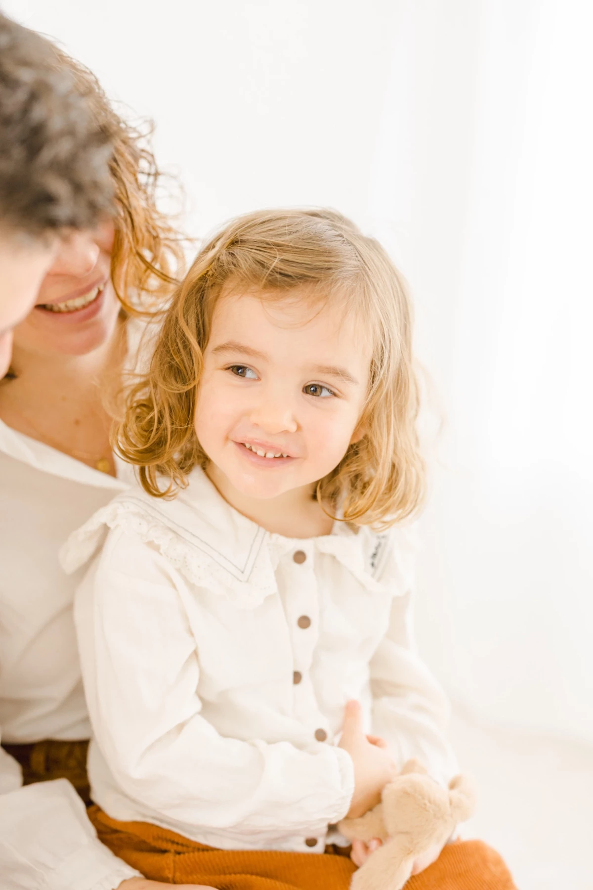 Mère et sa petite fille blonde en chemise blanche partageant un moment de complicité en studio