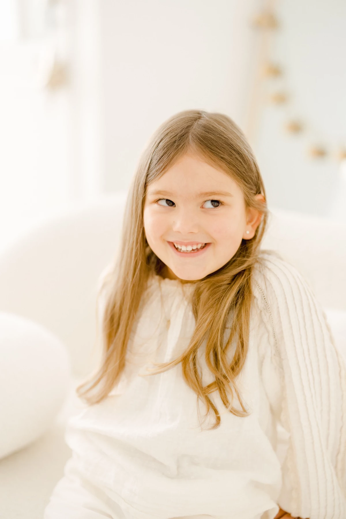 Jeune fille aux longs cheveux blonds souriante en pull blanc douillet dans un décor studio épuré