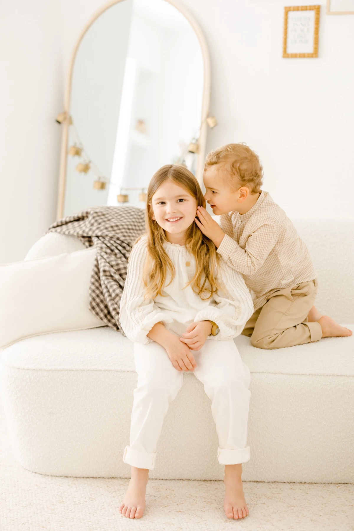 Deux enfants complices sur un lit blanc, un garçon chuchotant à l'oreille d'une fille souriante