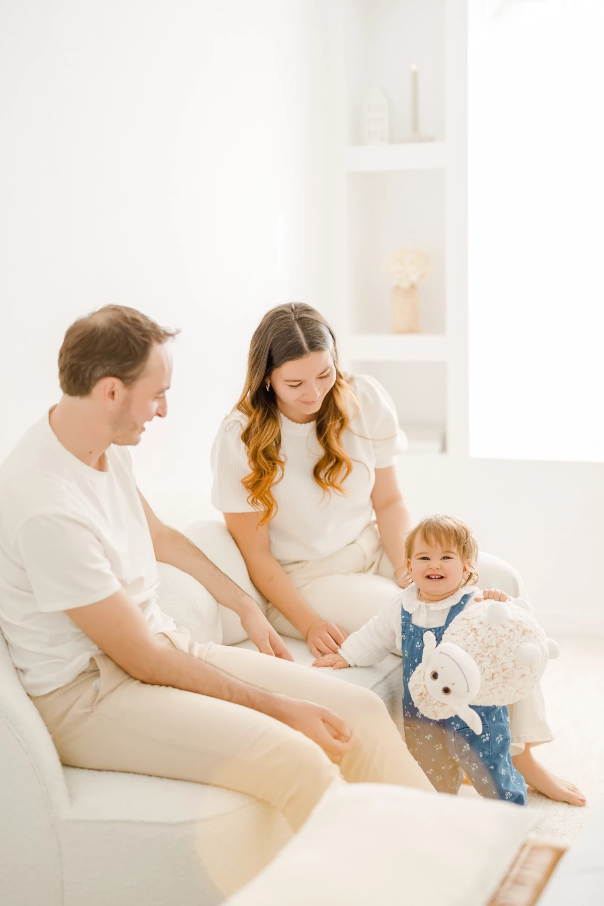Séance famille au studio : parents et petite fille partagent un moment de complicité avec une peluche dans une ambiance lumineuse
