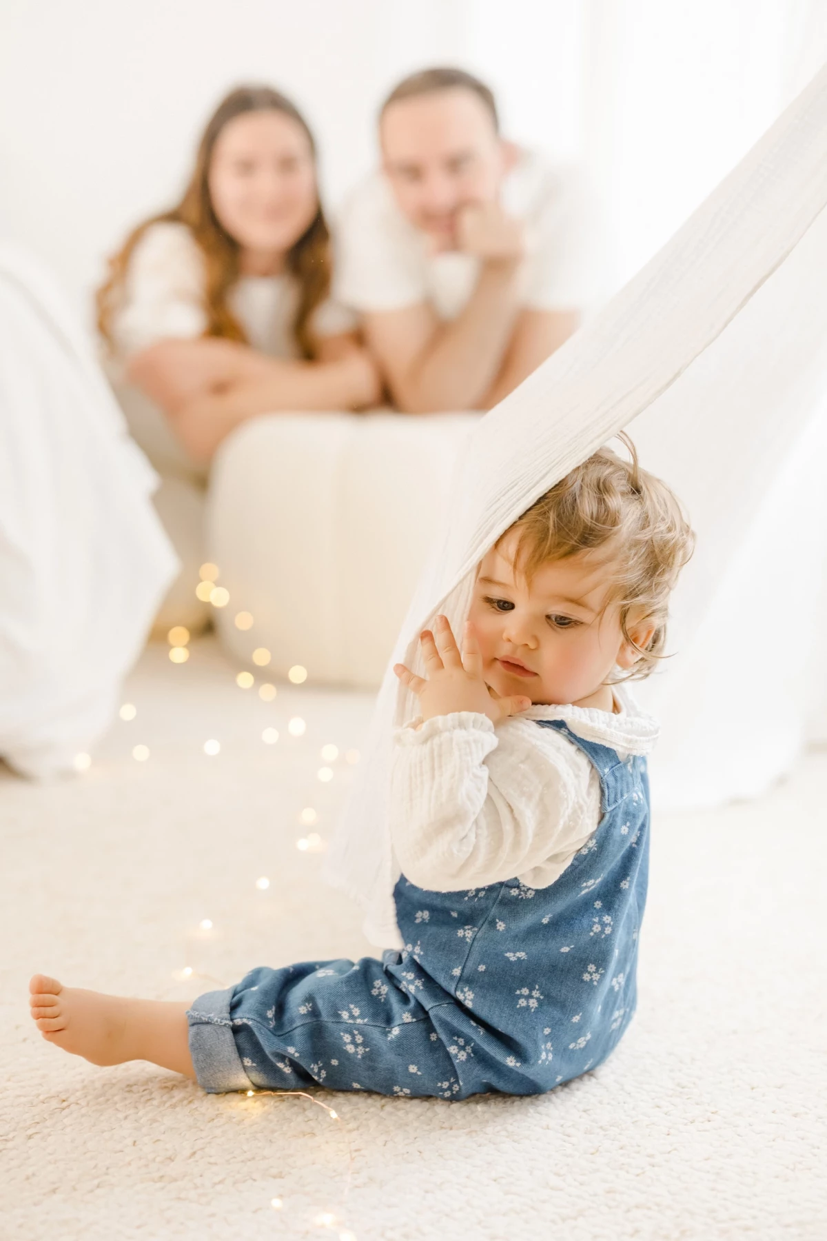Séance famille au studio : petite fille en salopette bleue assise devant ses parents, lumière douce et ambiance naturelle en Vendée