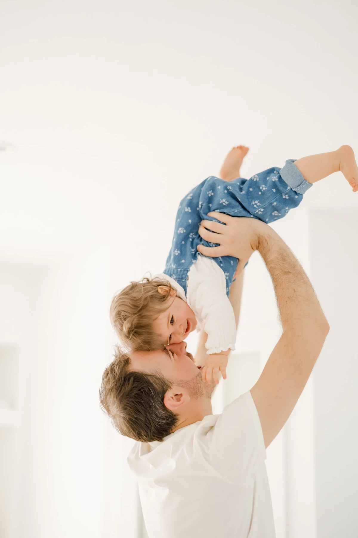 Séance famille en studio : père soulevant sa petite fille dans un moment de complicité joyeuse sur fond blanc lumineux