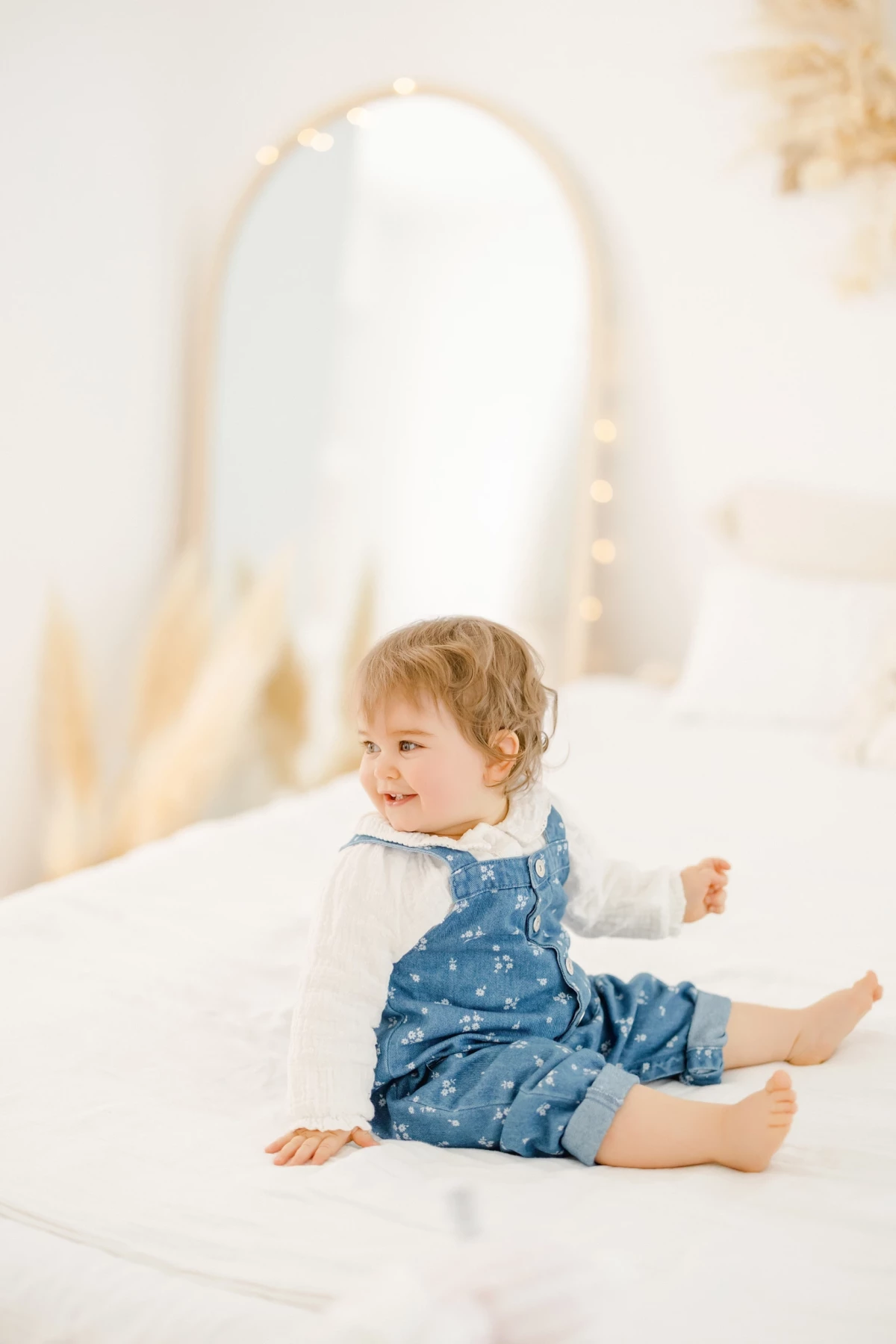 Séance famille en studio : petite fille assise sur un lit blanc, sourire complice et regard joyeux dans une ambiance lumineuse
