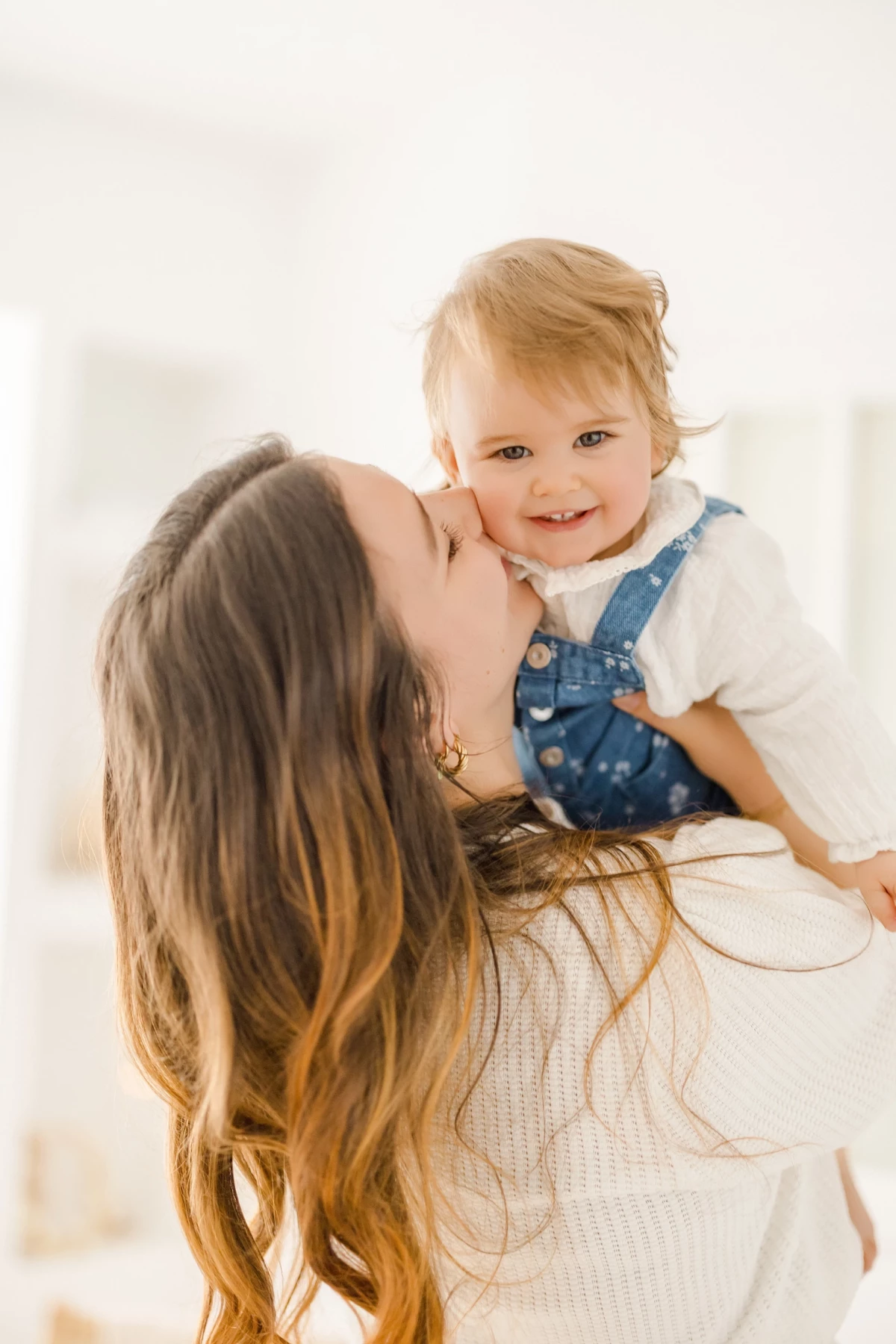Séance famille au studio : maman embrasse sa petite fille blonde souriante, moment de tendresse et complicité sur fond blanc