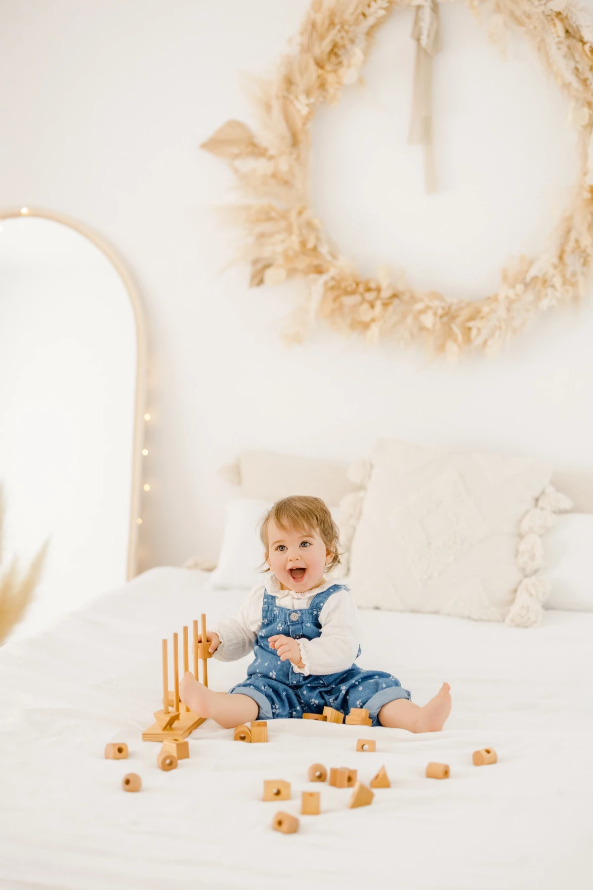 Séance famille en studio : petite fille souriante assise sur un lit blanc, entourée de jouets en bois dans une ambiance naturelle et douce