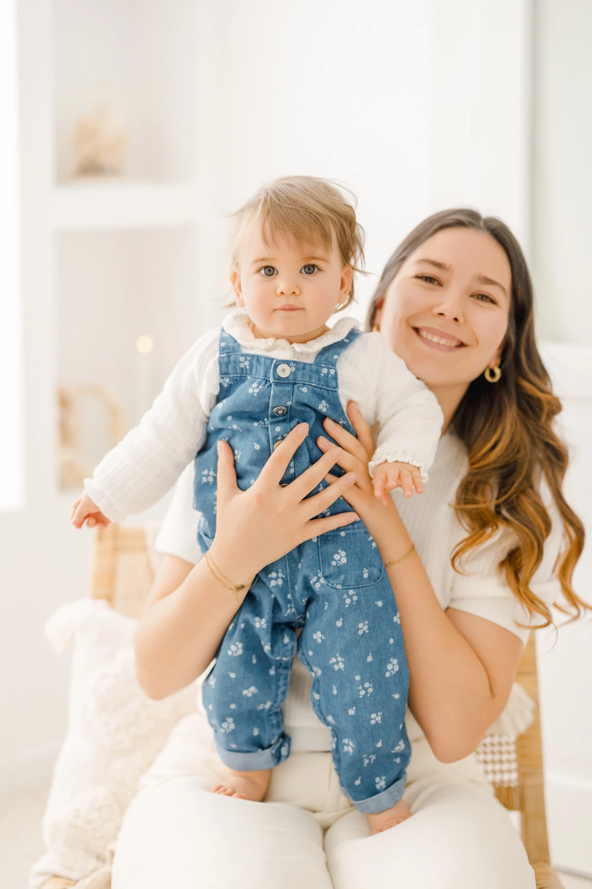 Séance famille au studio : maman souriante porte sa petite fille en salopette bleue, instant de complicité et tendresse maternelle