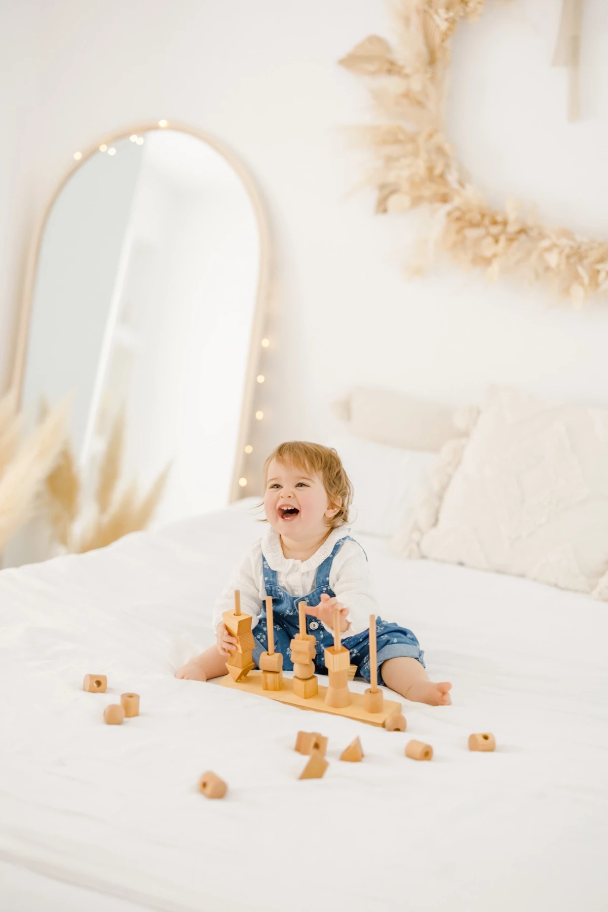 Séance famille en studio : petite fille blonde éclate de rire en jouant avec des cubes en bois sur un lit blanc