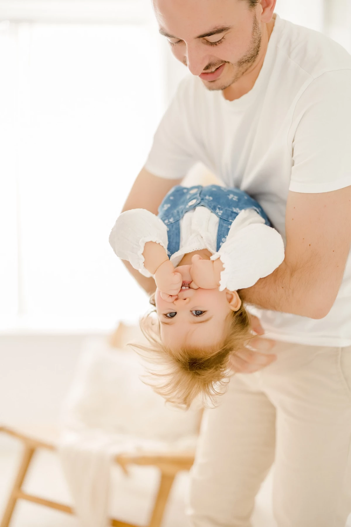 Séance famille au studio : papa joue avec sa petite fille en la retournant, moment de complicité joyeuse dans une lumière douce