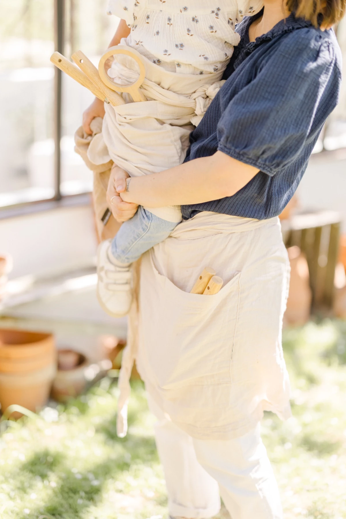 Séance famille en extérieur : maman porte son bébé dans un porte-bébé en toile beige, instant de douceur printanière dans une verrière lumineuse