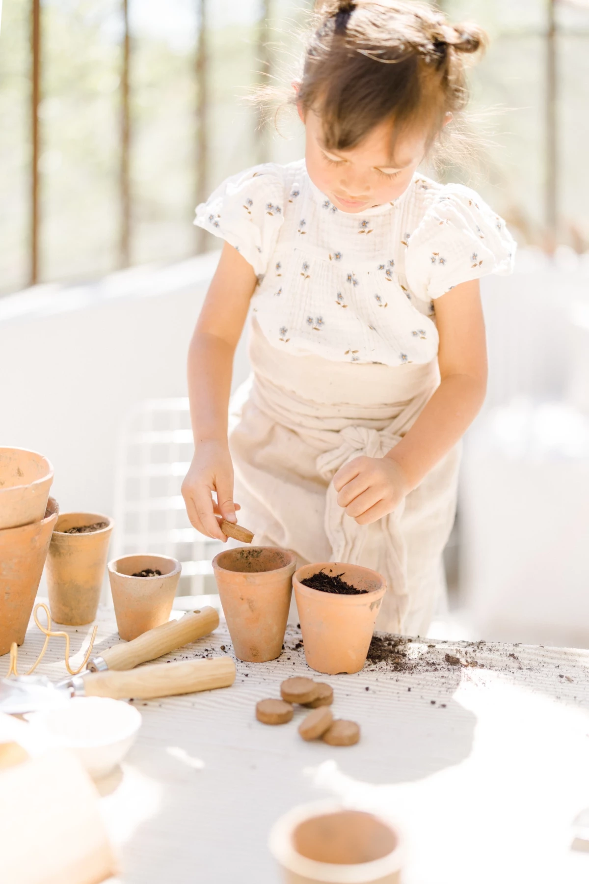Séance famille en verrière : petite fille concentrée plante des graines dans des pots en terre cuite, lumière naturelle printanière