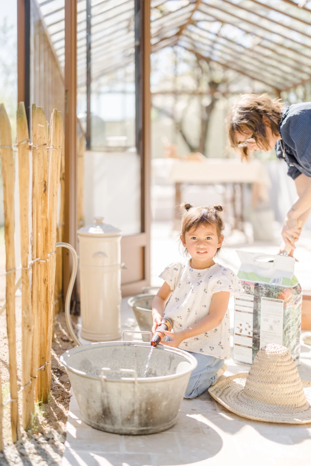 Séance famille printemps : fillette souriante joue avec l'eau dans une verrière lumineuse, complicité mère-fille authentique