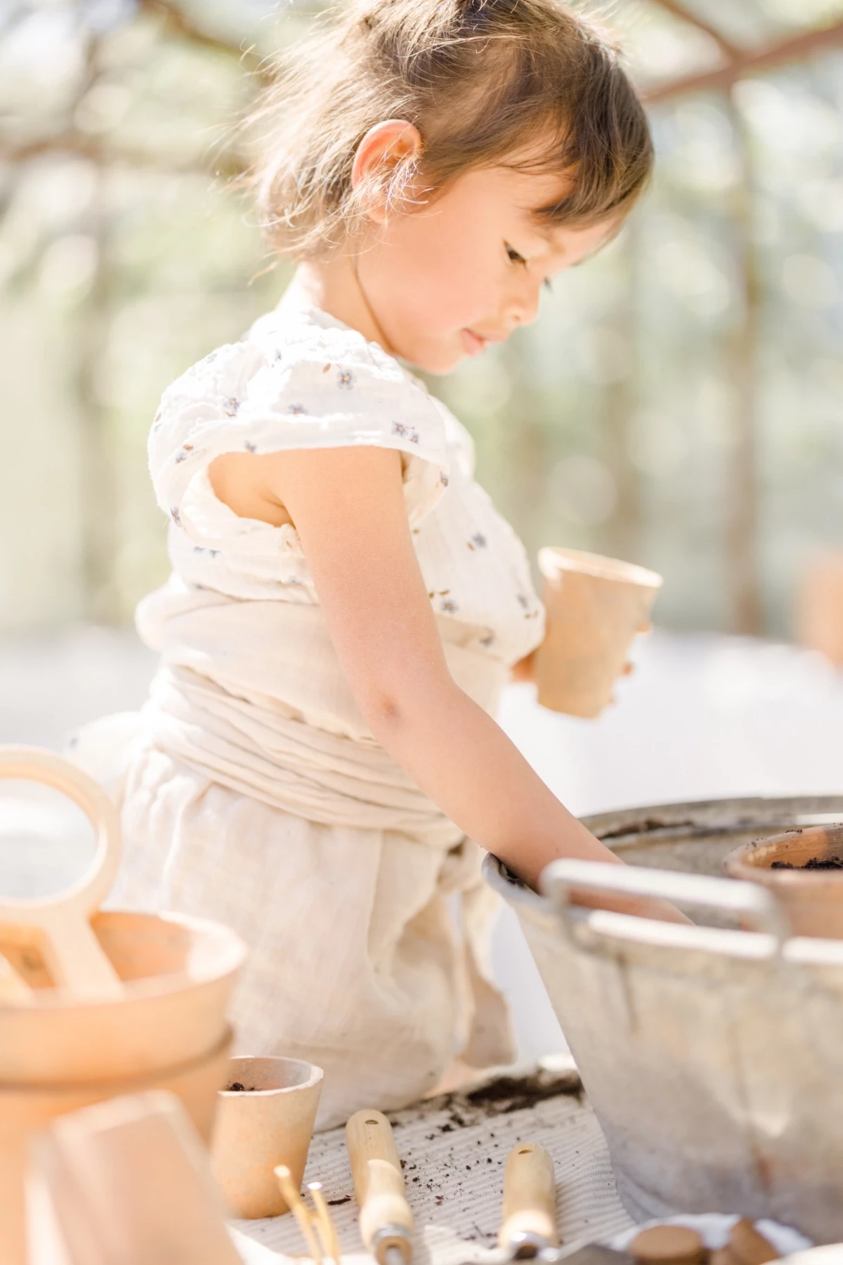 Séance famille en verrière : fillette concentrée lors d'un atelier jardinage, lumière printanière douce et naturelle en Vendée