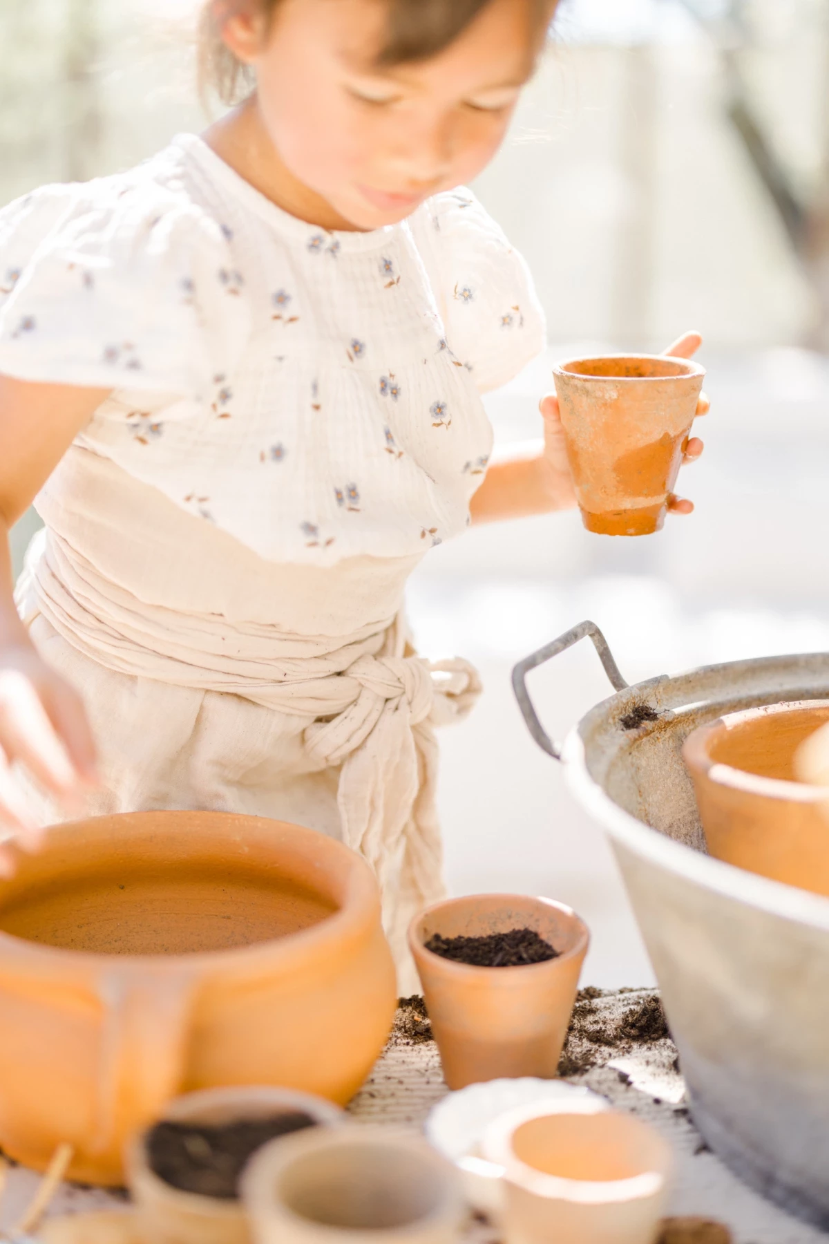 Séance famille printemps : petite fille joue au jardinage avec des pots en terre cuite dans une verrière baignée de lumière naturelle