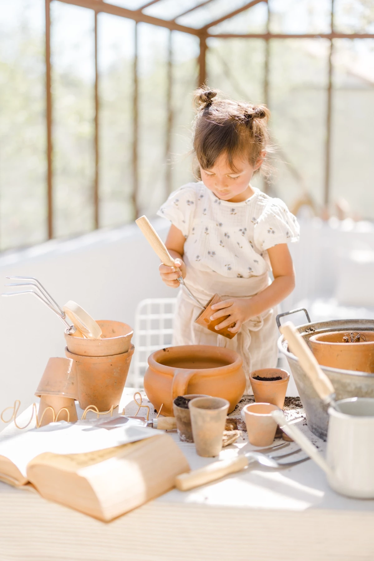 Séance famille en extérieur : fillette concentrée crée de la poterie dans une verrière lumineuse, moment d'éveil créatif printanier
