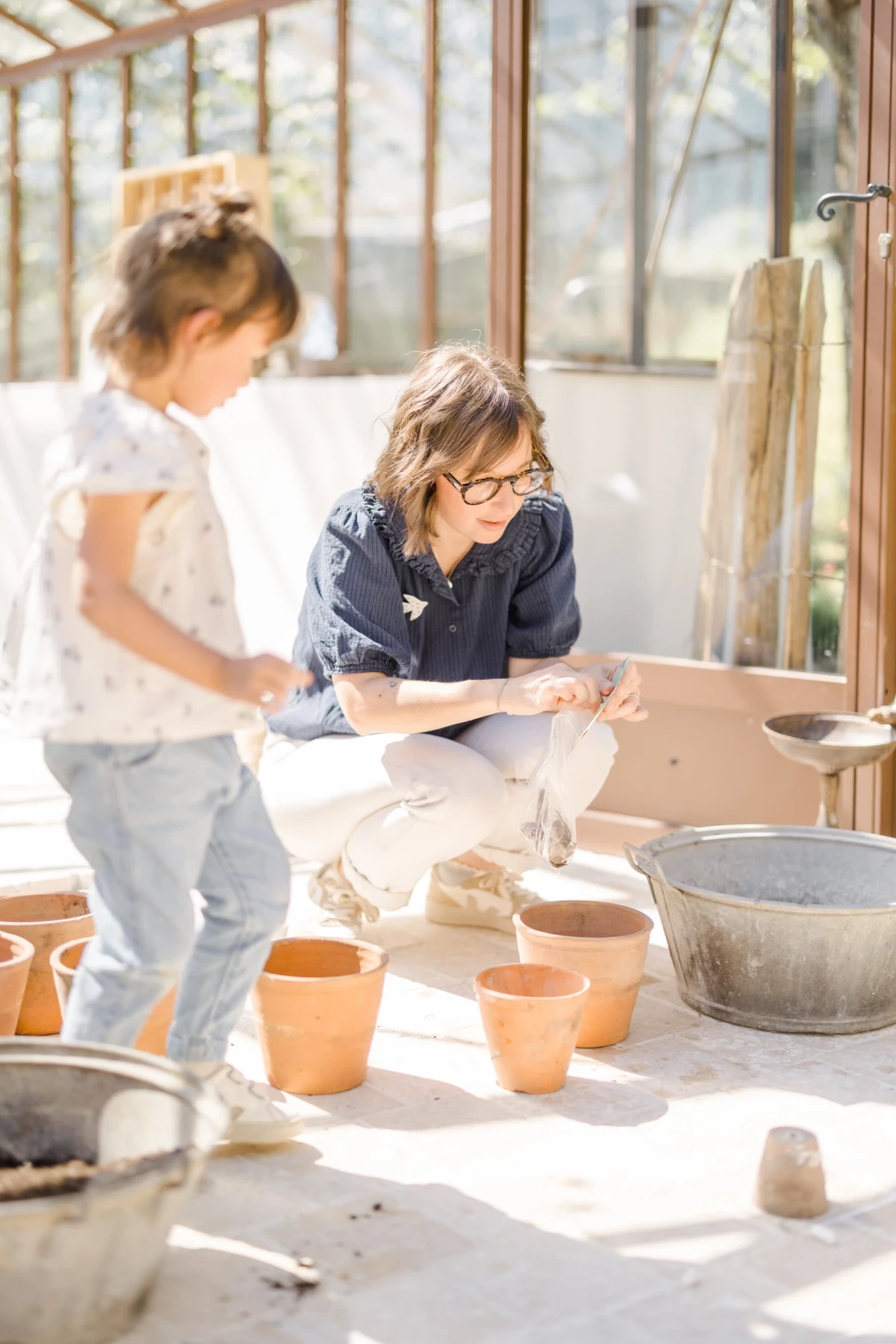 Séance famille en verrière : mère et fille partagent un moment de complicité autour du jardinage, lumière printanière douce