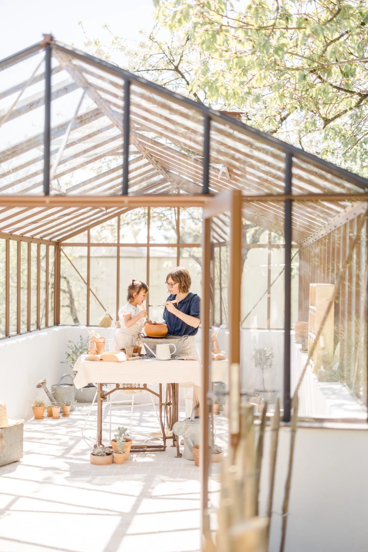 Séance photo famille en verrière : mère et fille partagent un moment complice autour du jardinage dans la lumière naturelle du printemps