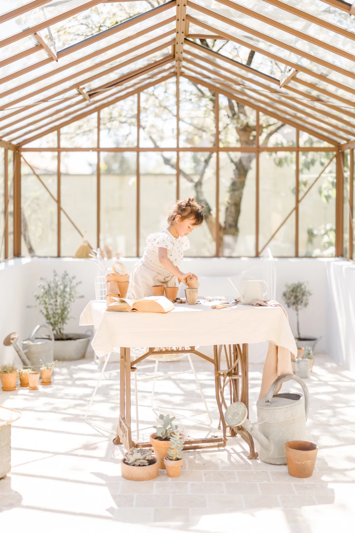 Séance famille printemps : fillette jardine dans verrière lumineuse, ambiance douce et naturelle en tons beiges et blancs