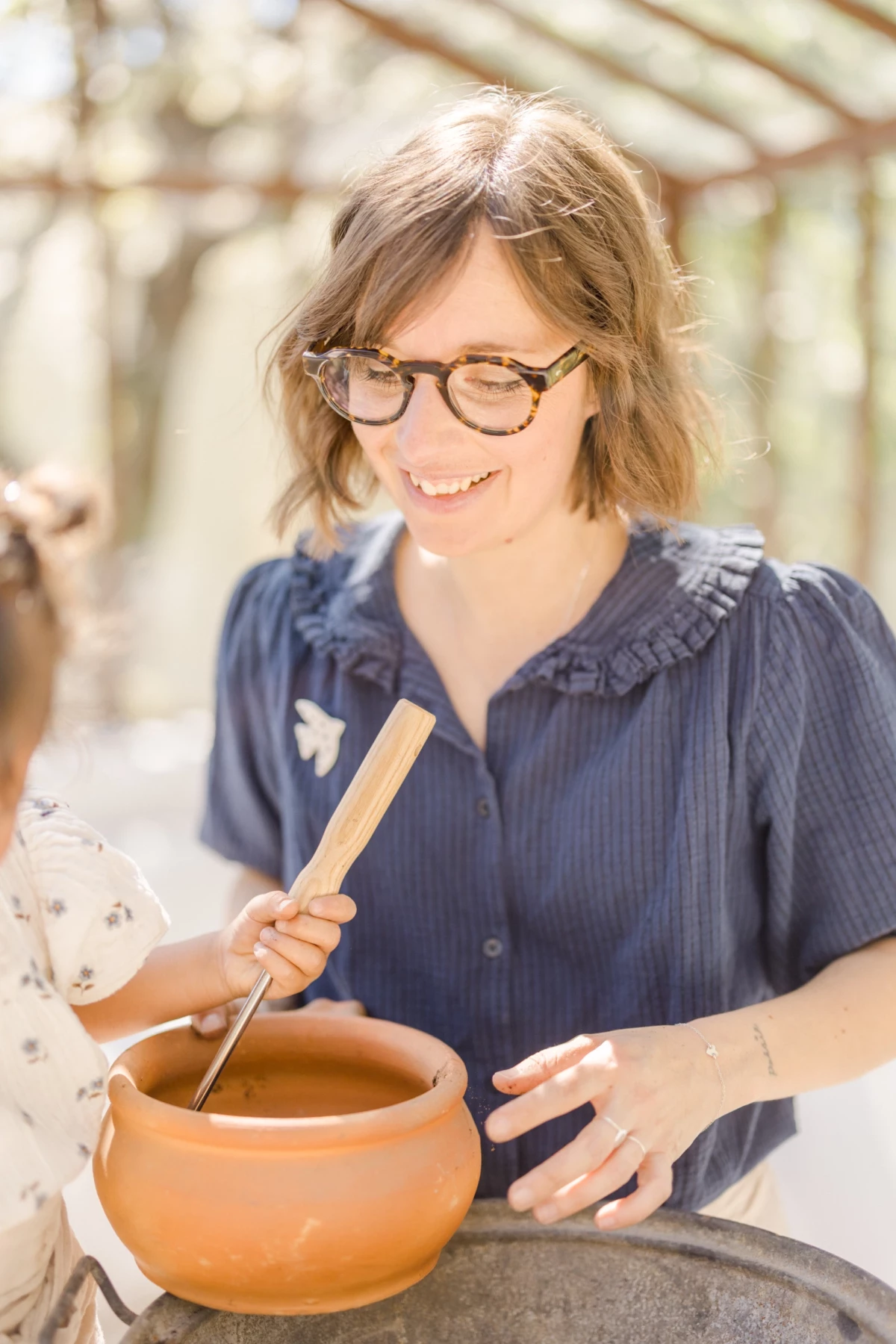 Séance famille en verrière : mère souriante partage un moment de jardinage avec sa fille, lumière naturelle printanière