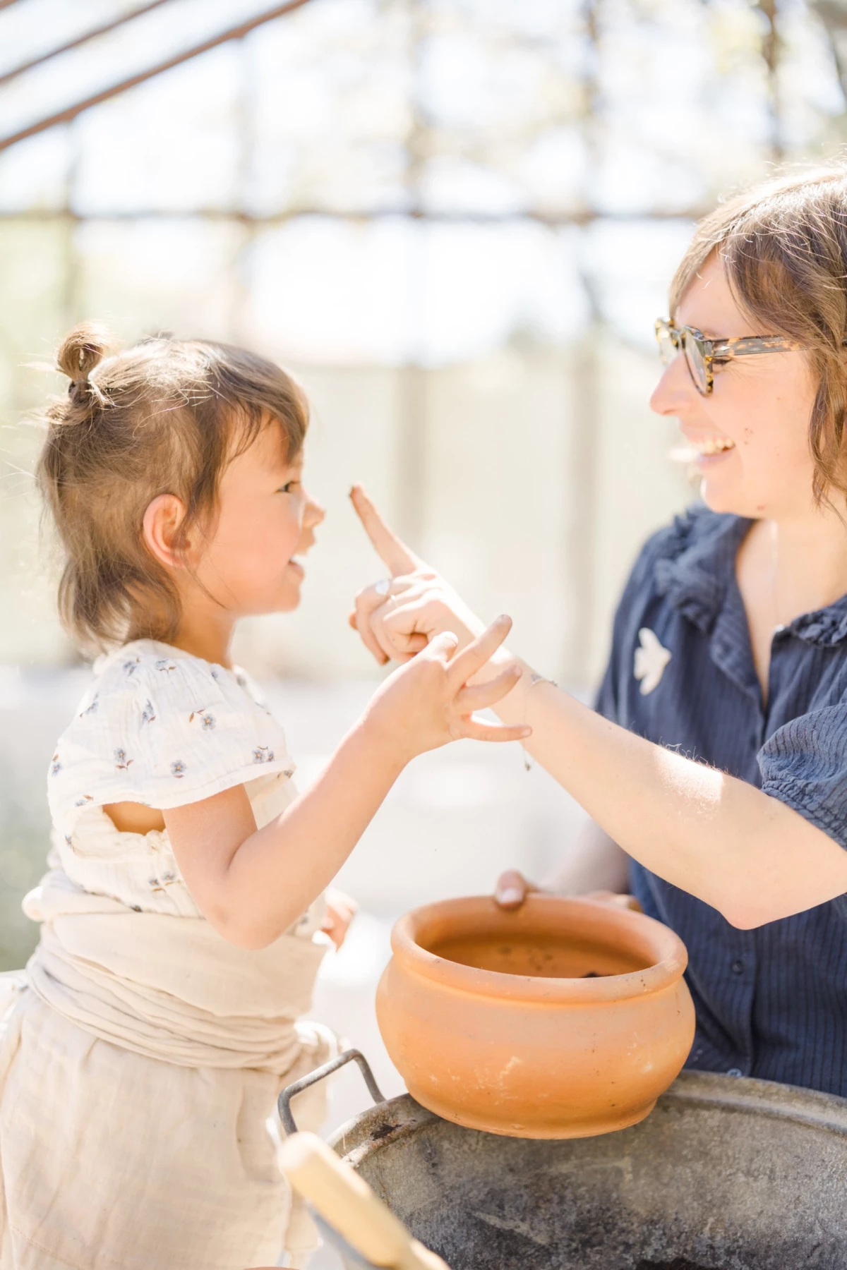 Séance famille en extérieur : mère et fille partagent un moment de complicité autour d'un pot en terre cuite, lumière douce de printemps