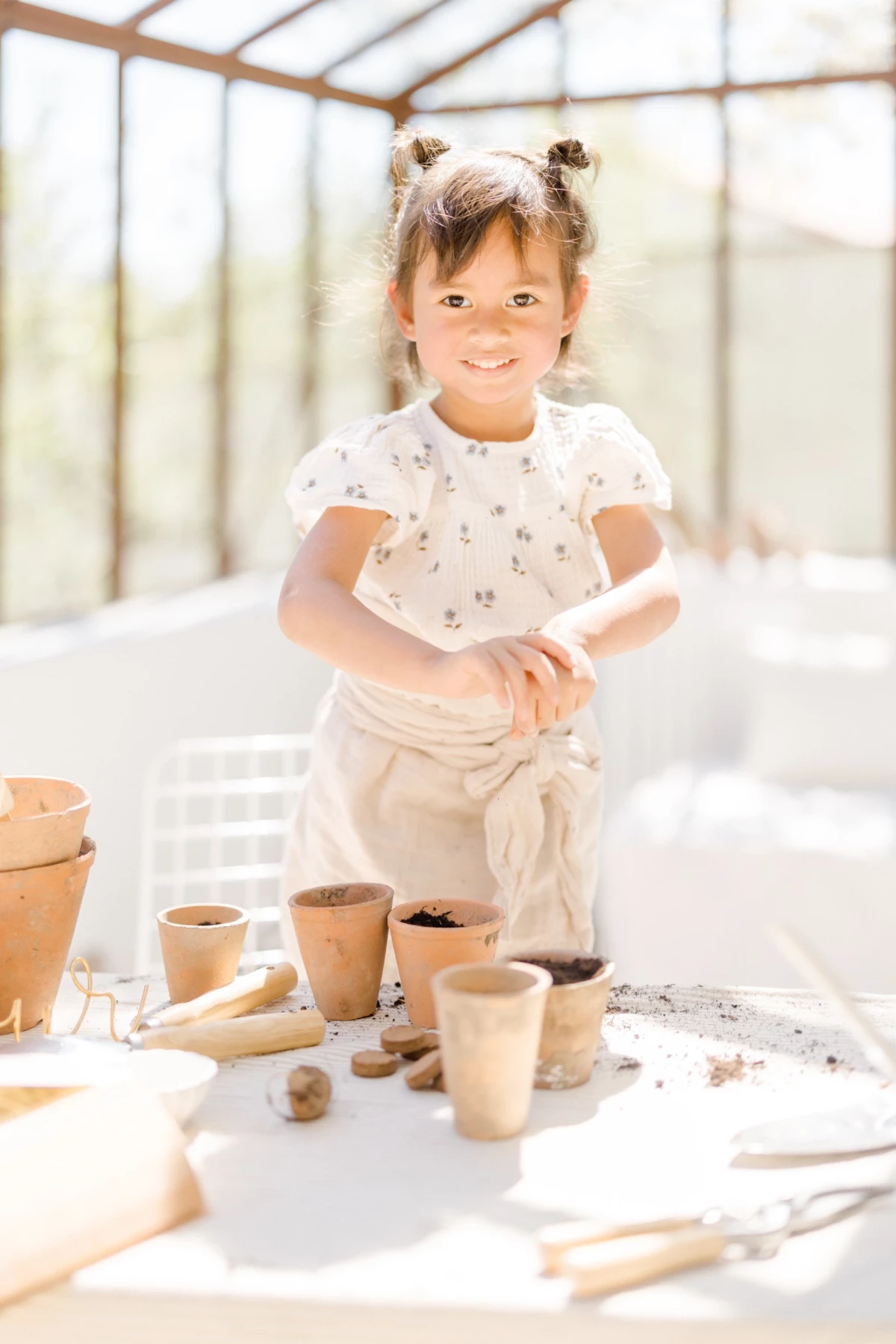 Séance famille printemps : petite fille souriante s'amuse avec des pots en terre dans une verrière lumineuse, ambiance douce et naturelle