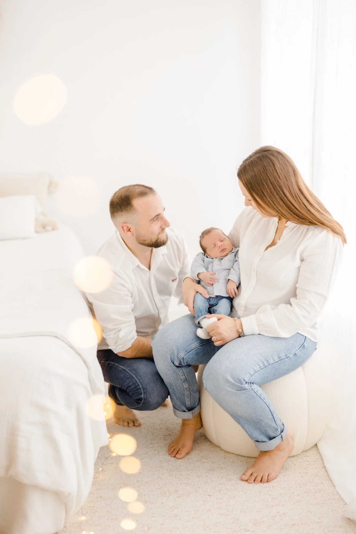 Séance nouveau-né en studio : jeunes parents assis face à face tenant leur bébé, ambiance douce et lumineuse