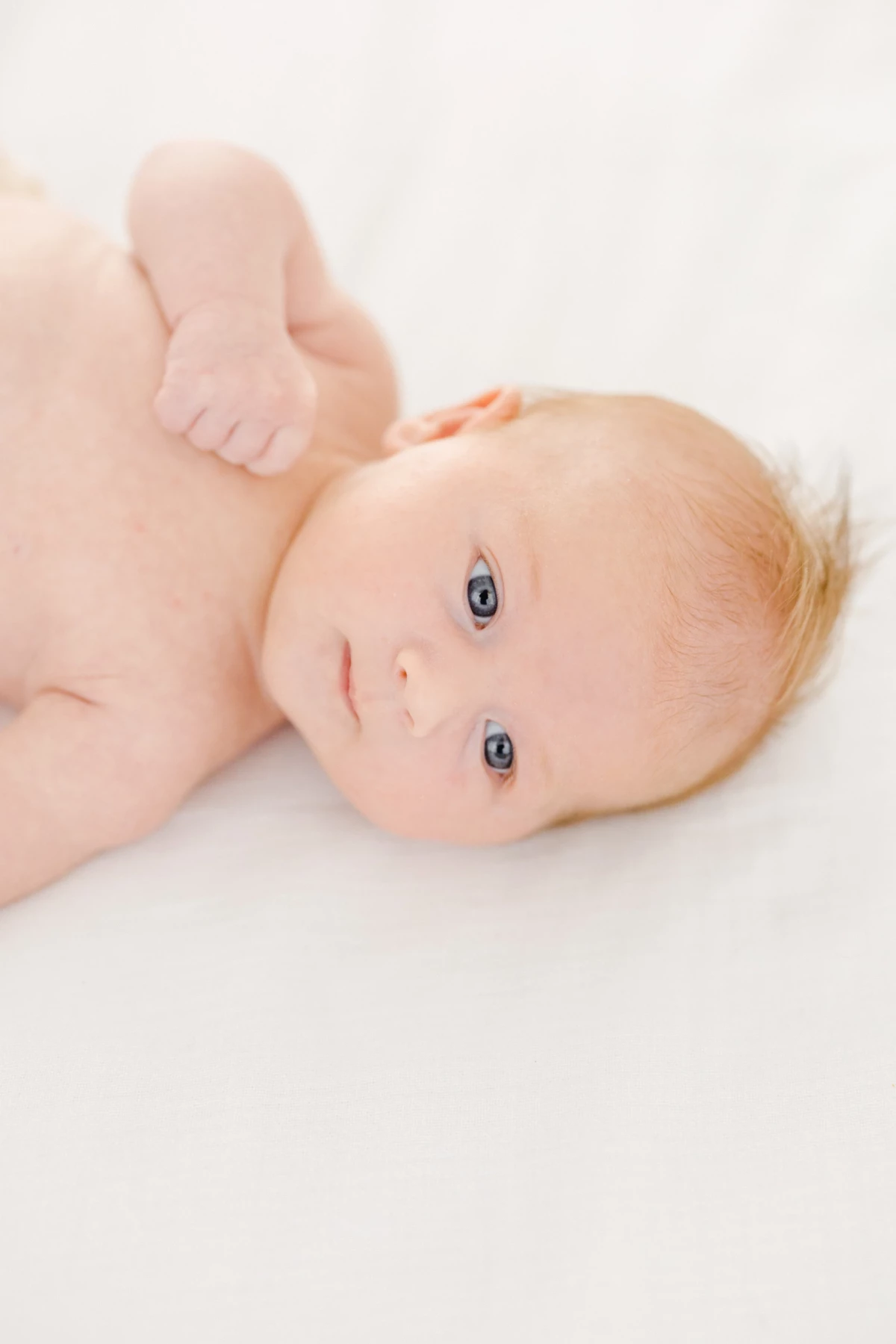 Séance nouveau-né au studio : bébé aux yeux bleus allongé sur fond blanc regarde la caméra avec tendresse et innocence