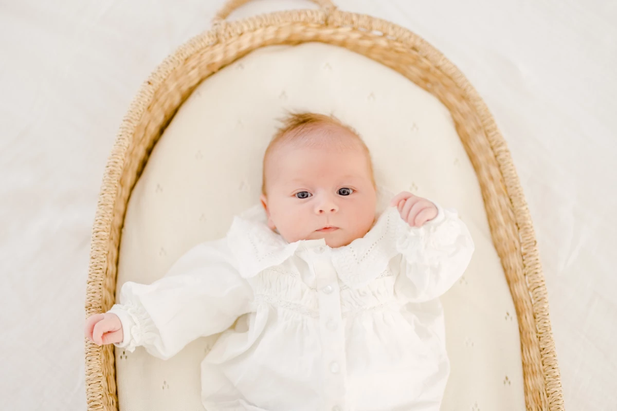 Séance naissance en studio : nouveau-né vêtu de blanc repose paisiblement dans un berceau en osier, tendresse infinie
