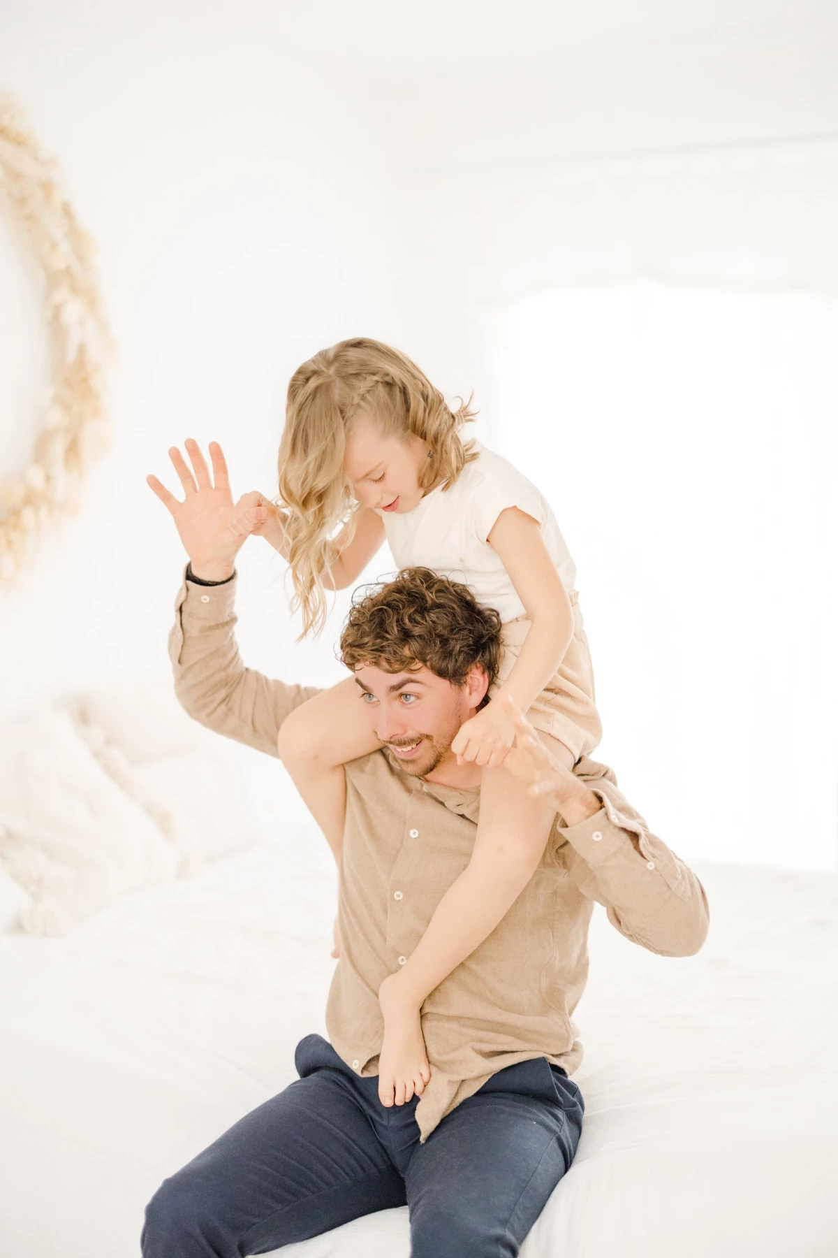 Séance famille en studio : père porte sa fille sur ses épaules dans une ambiance complice et lumineuse, décor épuré blanc et beige